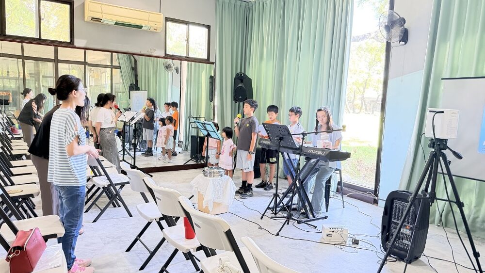 Several children singing into microphones at the front while a keyboard player accompanies them.