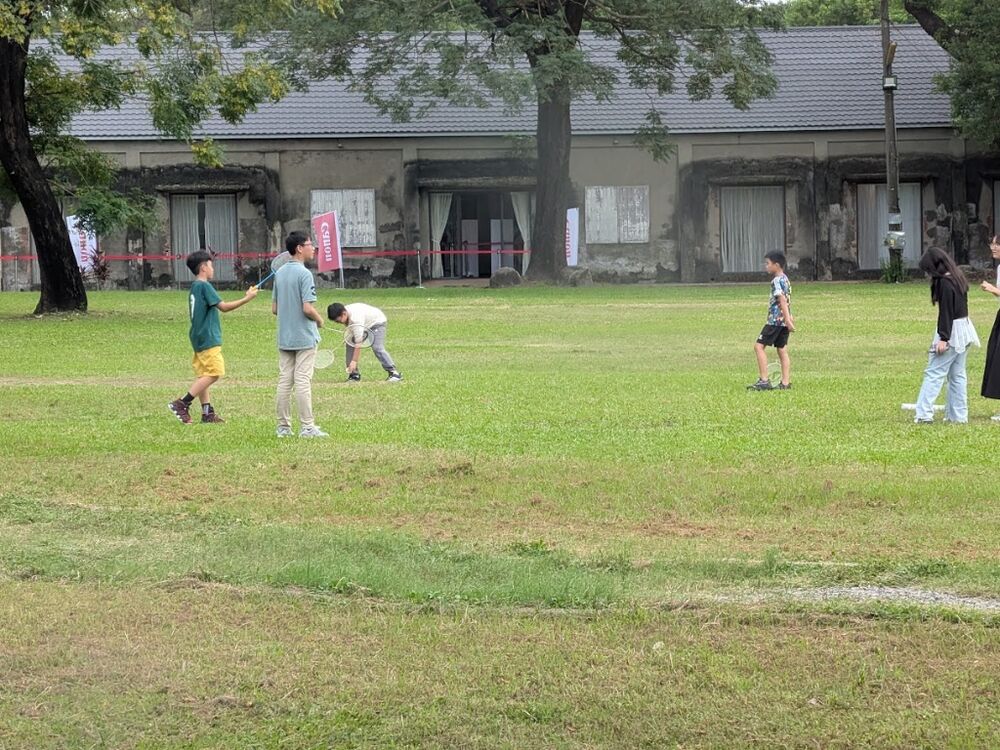 Adults and children playing badminton on the lawn, with Japanese-era sugar warehouse buildings in the background.