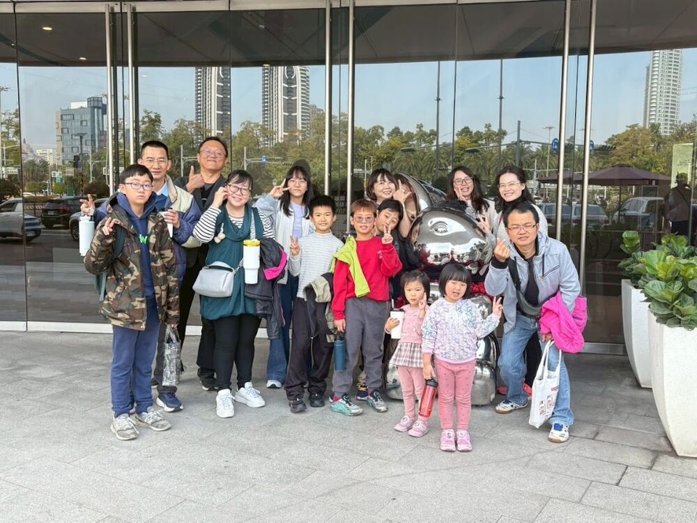 Small-group members and children posing in front of a building with a mirrored sphere sculpture.